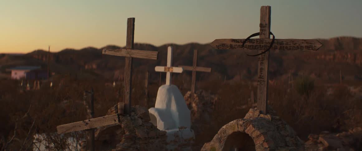 A cemetery scene at sunset with several wooden crosses and a white tombstone set against a backdrop of mountains and rural houses