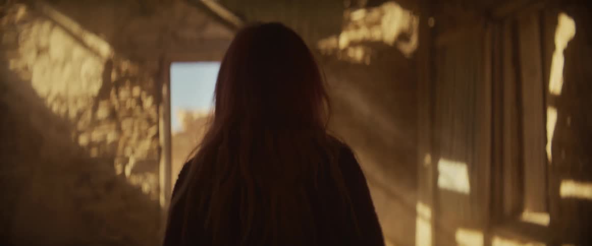 A woman with long wavy reddish hair walks through a dimly lit rustic room with textured walls illuminated by dappled sunlight streaming through holes and windows