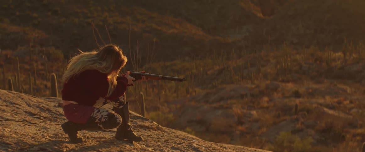A young woman or girl with long hair dressed in a red top and patterned pants kneels on a rocky desert terrain carefully aiming a rifle