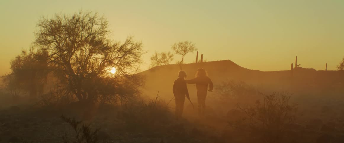 Two kids walk through a dusty desert landscape at sunset silhouetted against the glowing horizon