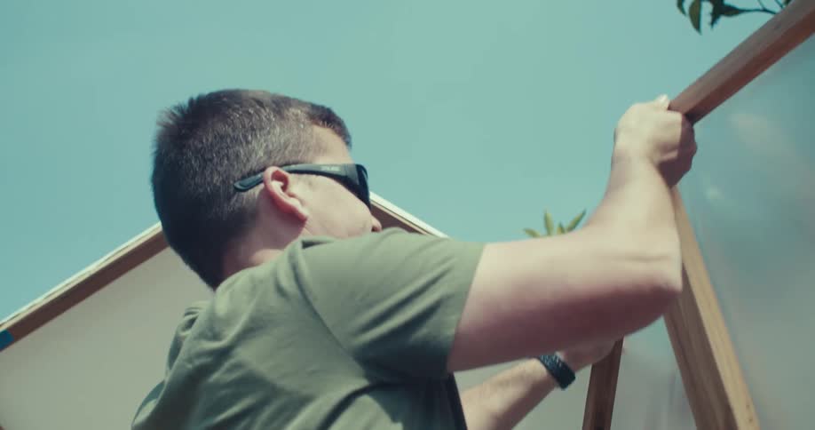 A person wearing sunglasses is working on the construction of a geodesic dome structure adjusting the plastic covering at the top