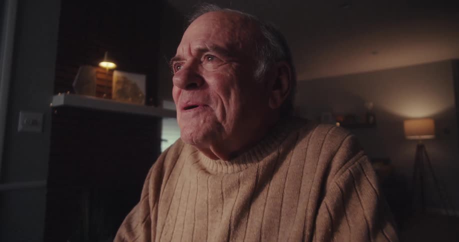 An elderly man sits at a table in a dimly lit living room smiling as he becomes lost in thought while working on his laptop which is adorned with a sticker