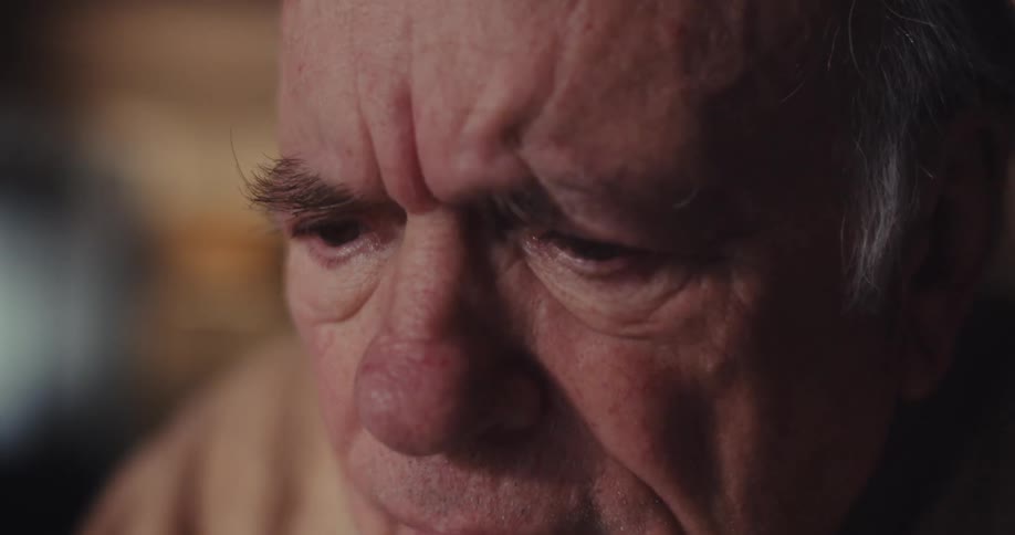 An elderly man sits at a table in a dimly lit living room smiling as he works on his laptop