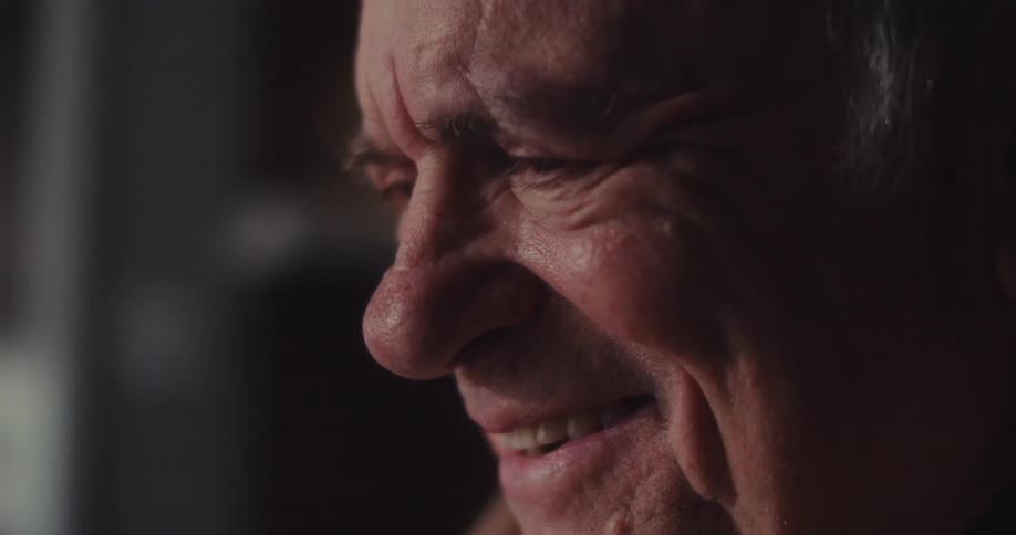 An elderly man sits at a table in a dimly lit living room smiling as he is working on his laptop