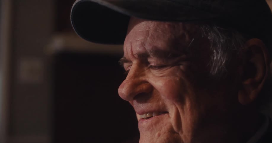 An elderly man wearing a dark cap with an american flag and the word veteran smiles in a dimly lit room