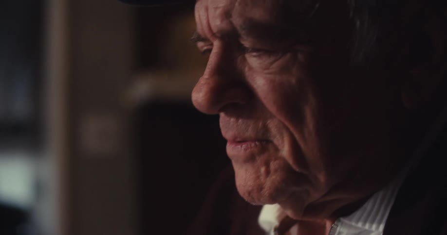 An elderly man sits at a table in a dimly lit living room focused as he works on his laptop