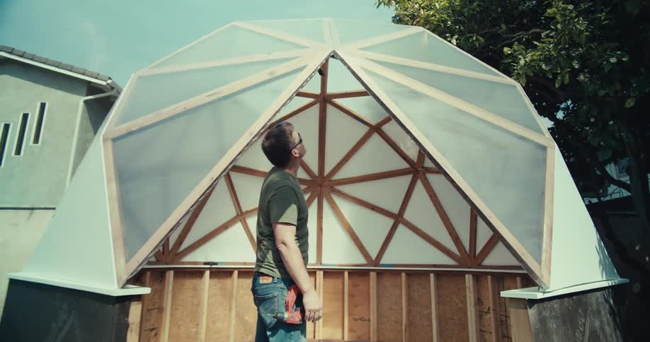 A person wearing sunglasses is working on the construction of a geodesic dome structure adjusting the plastic covering at the top