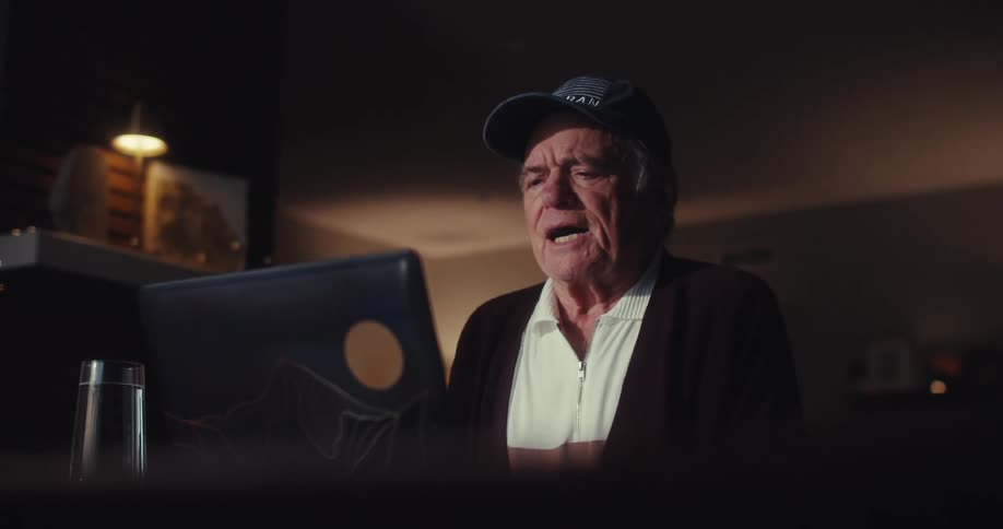 Elderly man sitting in front of a computer at home at night talks