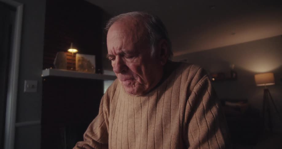 Elderly man frowns in thought before laughing while watching his computer at home at night