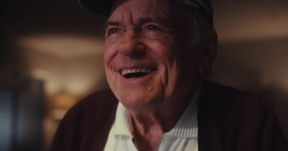 An elderly man in a room typing on his laptop and laughing