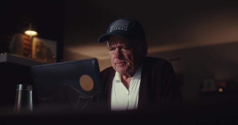 Elderly man sitting at home frowns while viewing a computer screen at night
