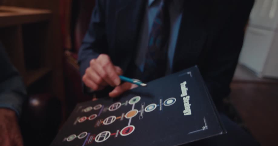 An elderly man is shown an advertisement by a man in a suit as they both sit in the living room area