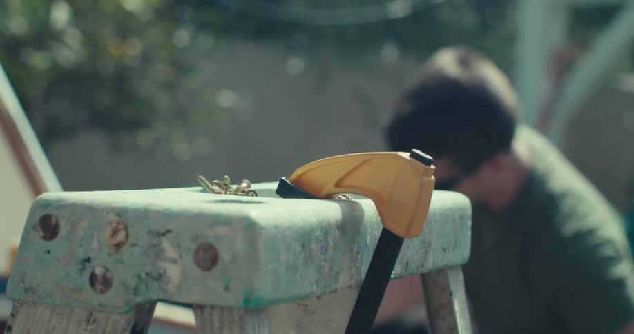 A closeup shows screws and a yellow clamp resting on a worn step stool with a man working blurred in the sunny background outdoors