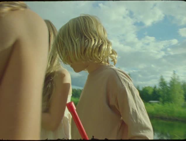 Three kidstwo girls and one boystand beside a lake on a sunny day laughing and smiling as they look at the palm of one of the girls