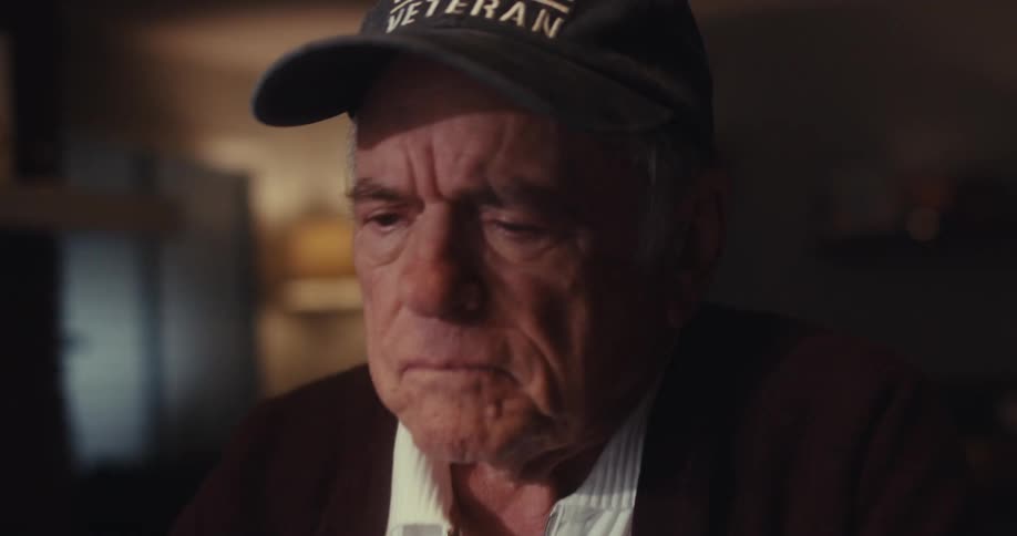 A closeup of an elderly man wearing a hat his expression thoughtful as he sits in a softly lit room