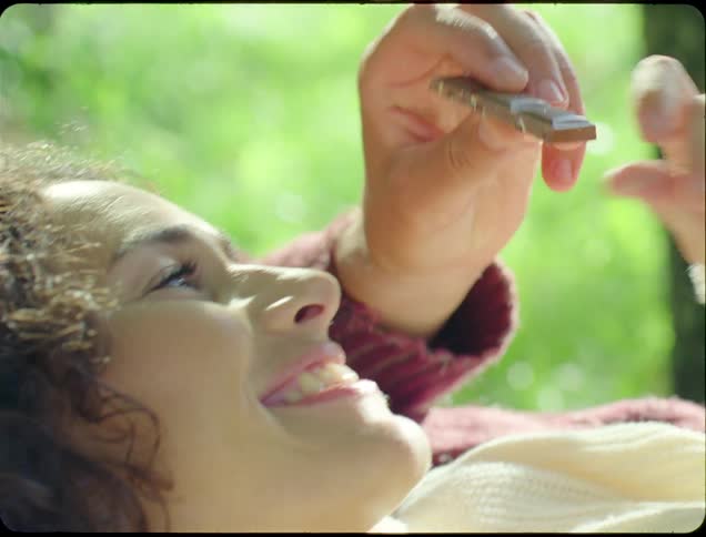 The face of a smiling woman who is looking at a piece of chocolate that is in someone elses hand