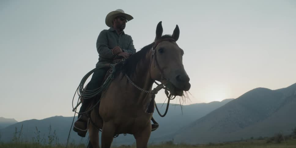 A person wearing a cowboy hat sits atop a horse that is grazing in a field with mountains in the distance under a morning sky