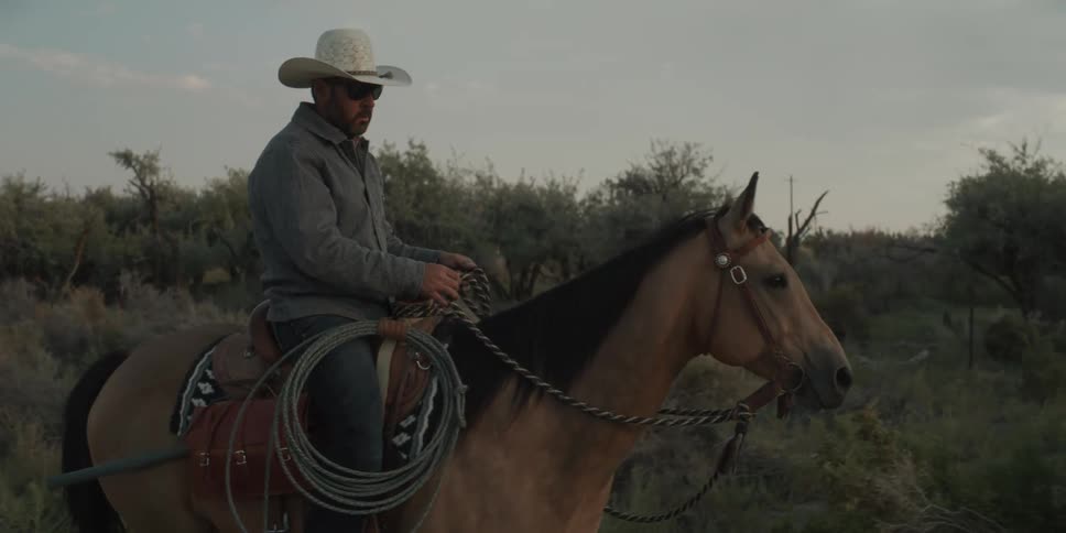 A cowboy wearing a hat and sunglasses rides his horse along a dusty path through brush holding the reins and carrying a coiled rope
