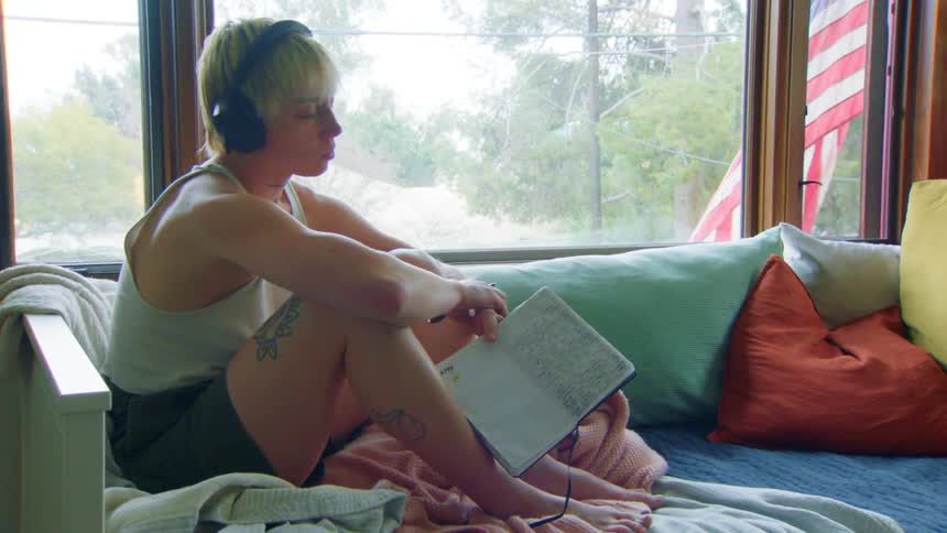 A woman with short blond hair wearing headphones sits comfortably by a window with a notebook and pen closing their eyes and jamming out to the music they are listening to