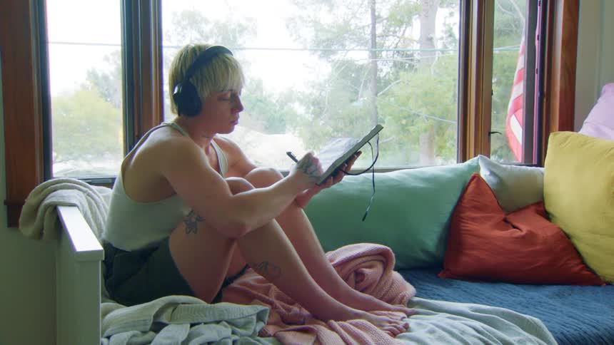 Blonde man sits on a couch near the window and writes in a journal while listening to music