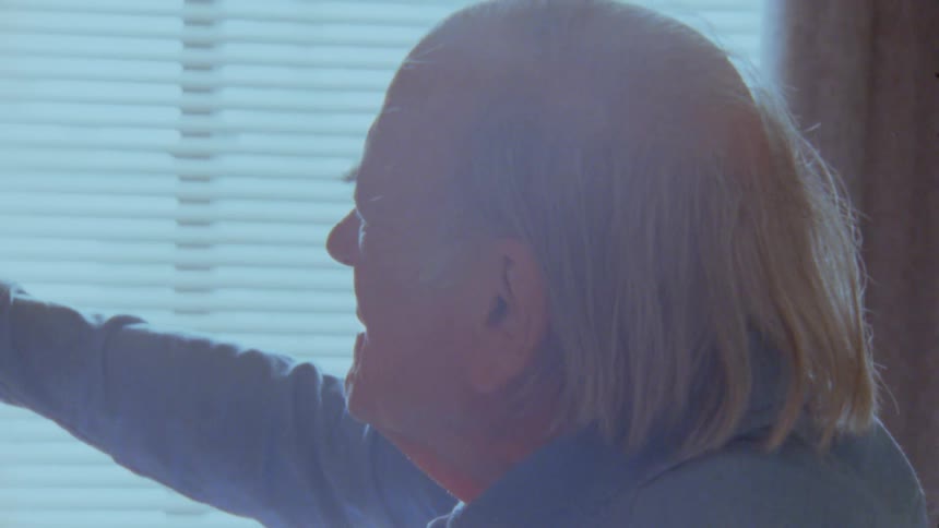 An elderly man is shown indoors pointing and using a remote control silhouetted against a window with blinds