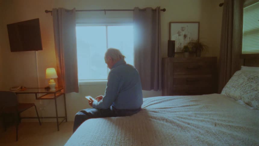 An elderly man sitting on a bed looking at a cell phone near a window