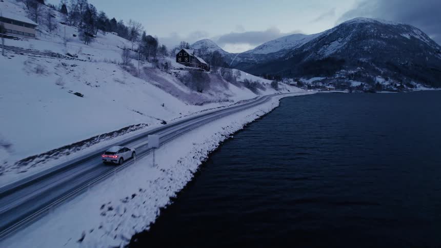 A vehicle driving on a road next to a snow covered hill and on the other side there is a huge lake with dark water