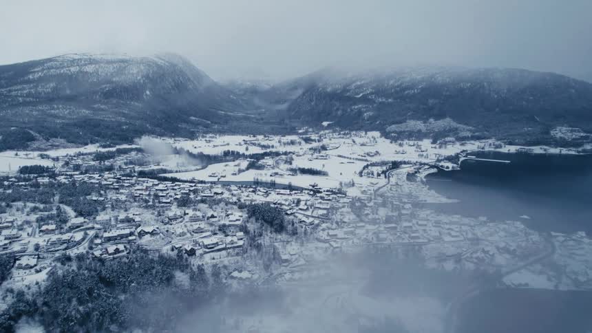 An expansive aerial view reveals a snowcovered coastal town nestled between mountains and a large body of water with wispy clouds drifting across the scene