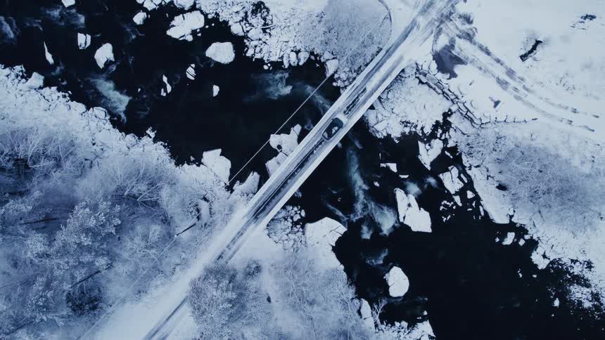 A topdown aerial view shows a car driving across a snowcovered bridge spanning a partially frozen river in a winter landscape