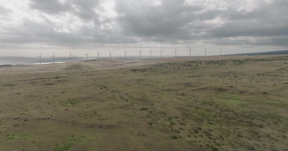 A windmill stands with a series of wind turbines turning beyond a grassland on a cloudy day