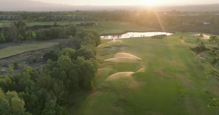 At sunrise on a golf course sprinklers spray water across the grass field as the sun rises behind a series of trees and vegetation