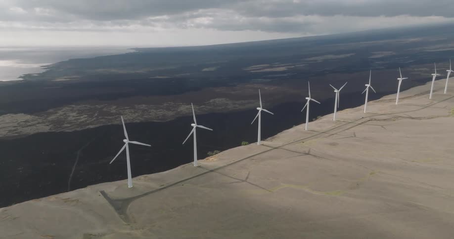 A series of windmills stand on a hill near a cliff rotating with an ocean view on the other side of the cliff at midday