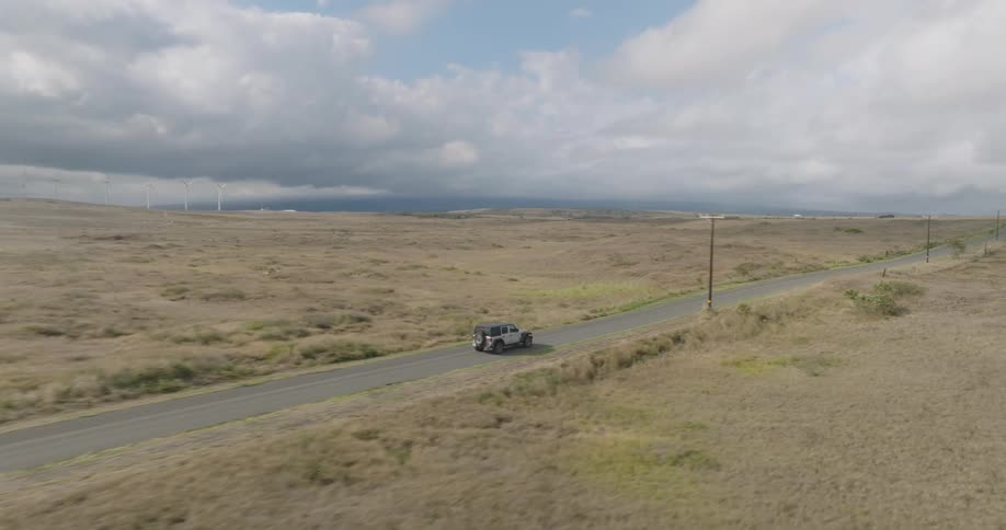 Vehicle travels along a rural highway passing wind turbines and pasture land on a cloudy day