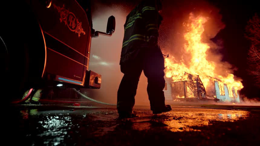Slow motion of fireman walking towards a house engulfed in flames at night