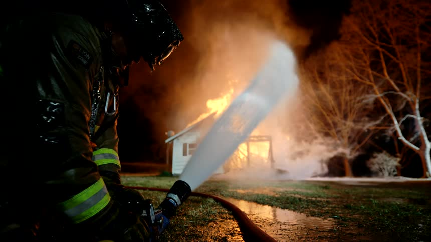 A firefighter holds a hose directing water toward a burning suburban home surrounded by trees at night