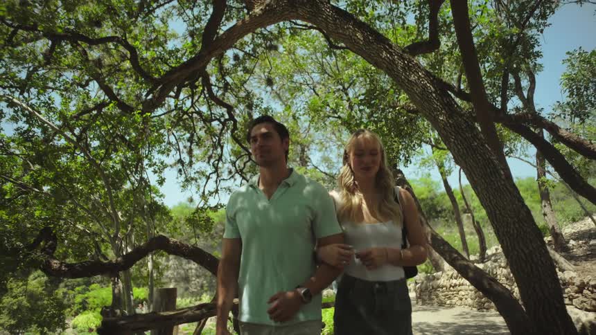 A man and woman are walking arminarm along a path with rocky walls and lush foliage under a bright sky