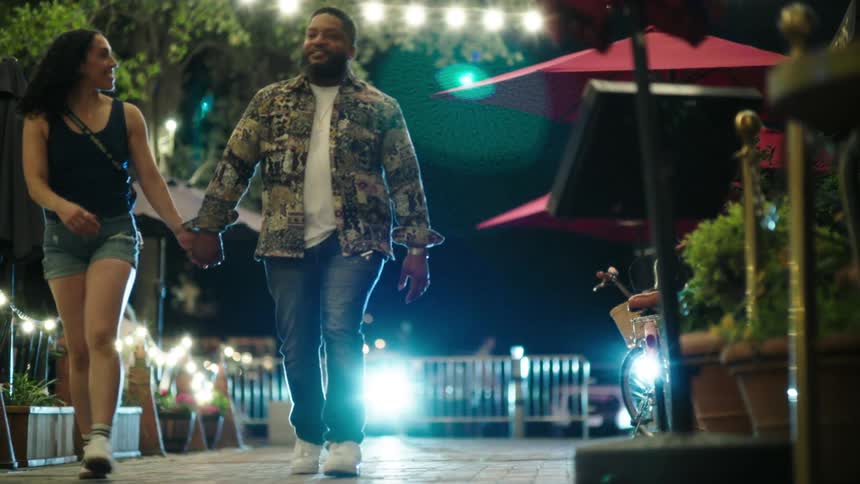 A happy couple walks hand in hand through a lit outdoor dining area at night