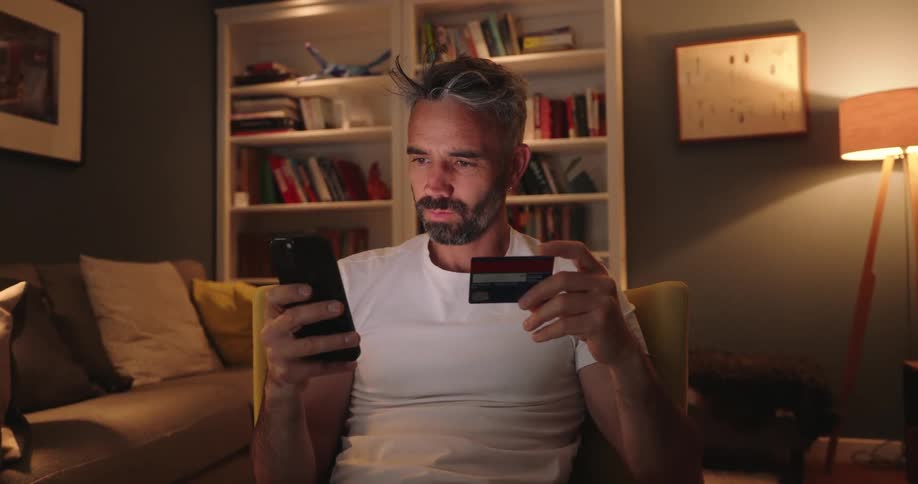 A man sitting on a chair in the living room using his phone and holding a credit card