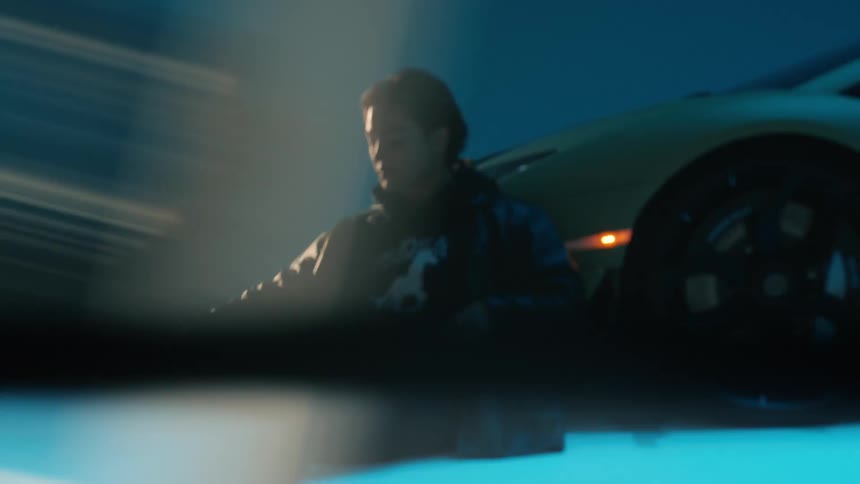 A man sits on the floor inside a garage in front of a sports car wearing a jacket as blue light flickers around him