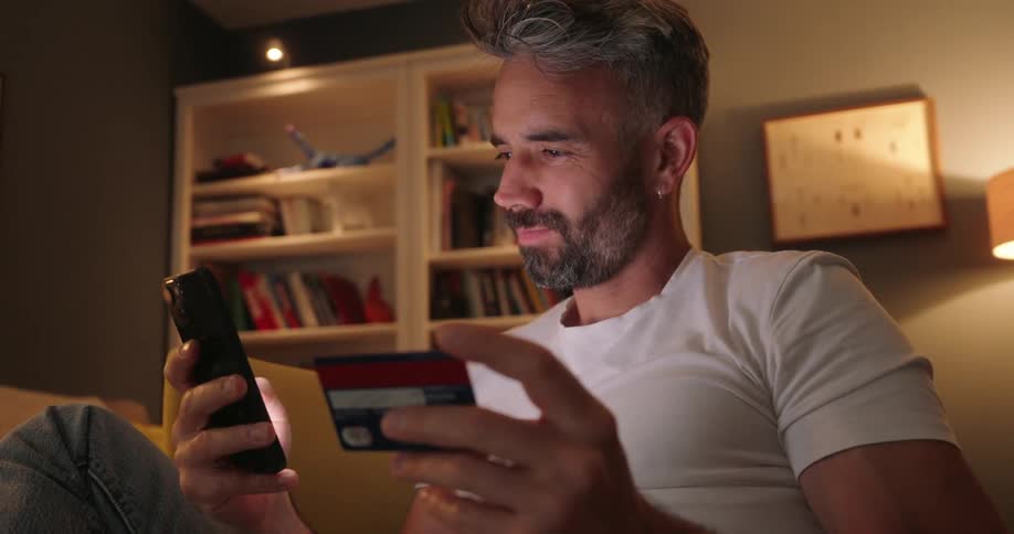 Man seated indoor types mobile phone while holding a credit card at night