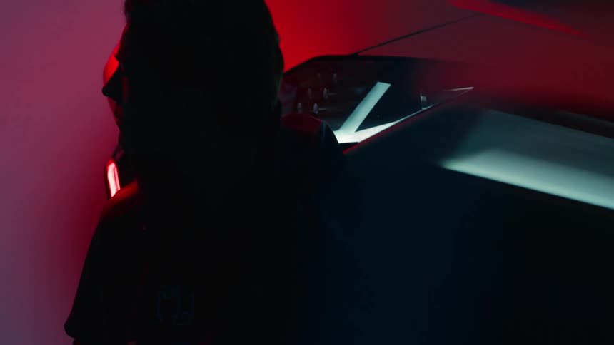 A man sits in front of a sports car inside a garage at night with the cars headlights on and blue light flickering in the room