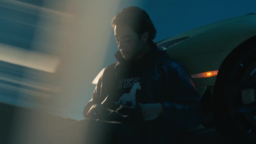 A man wearing a jacket sits in front of his car near the headlamp at night