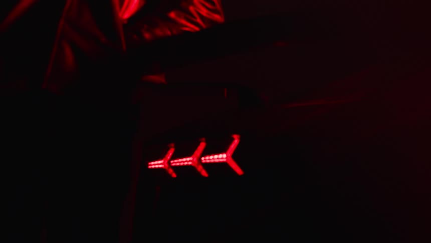 A man wearing a jacket stands behind a sports car with the taillights on inside a dark environment with flickering blue light