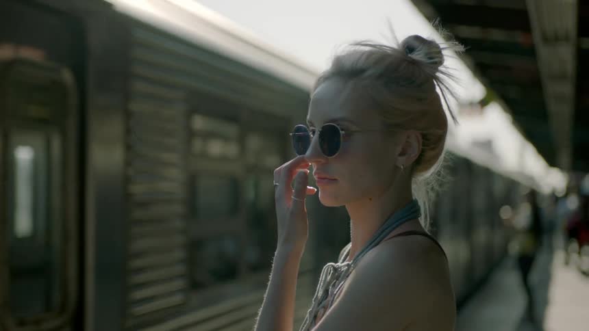 Blond girl wearing sunglasses stands while a train comes into the station