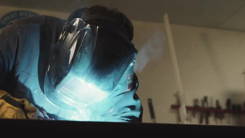 A person wearing a welding mask works intensely with bright sparks flying in a workshop