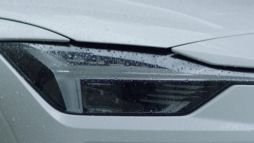 A closeup shot shows a cars headlight blinking while raindrops cover the vehicles surface