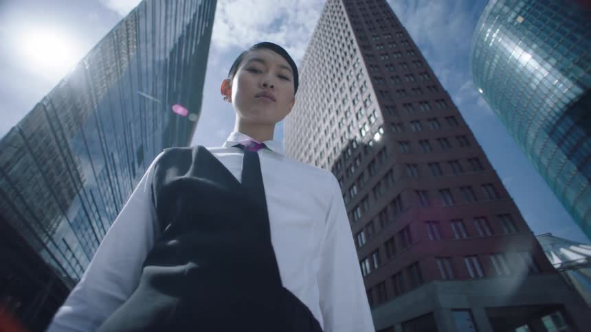 A confident person in formal wear poses beneath skyscrapers under a bright blue sky