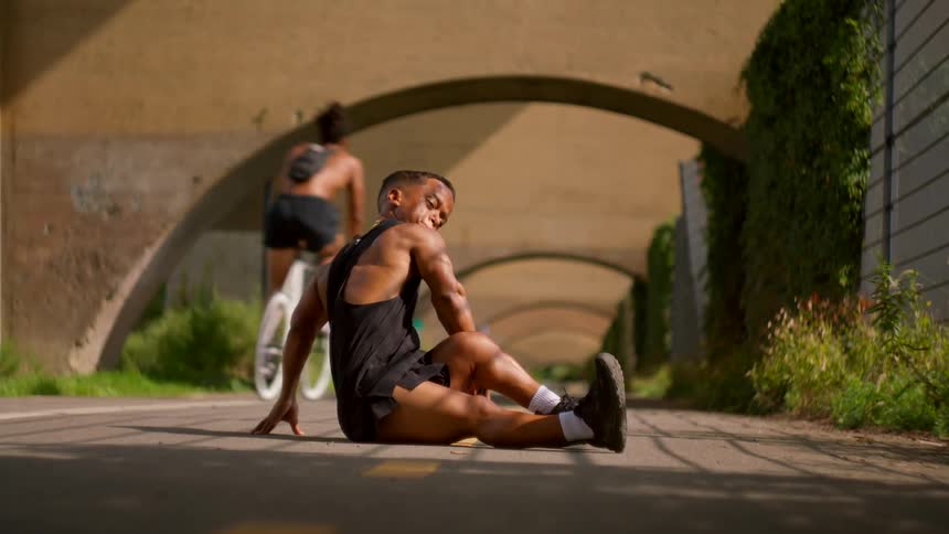 A young black man stretches on a path as a cyclist passes under arched bridges