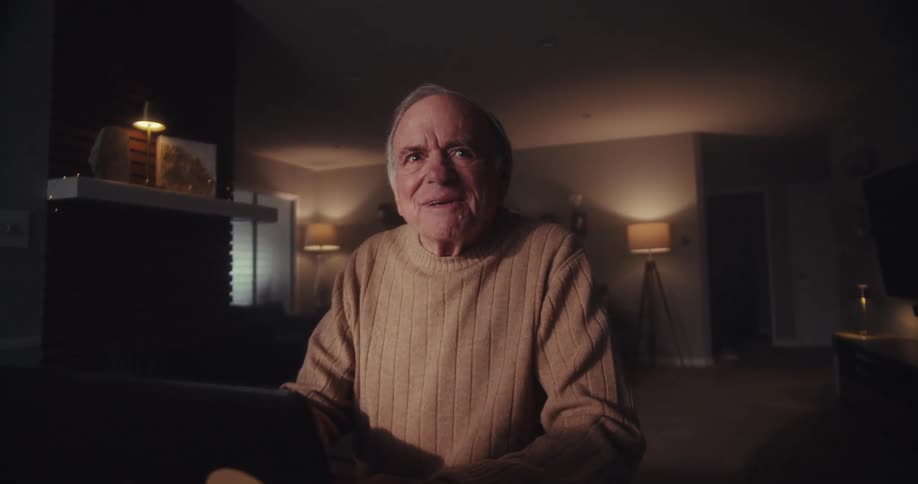 An elderly man smiles while video chatting on his laptop in a cozy dimly lit room
