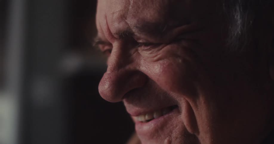 An elderly man laughs joyfully while video chatting on his laptop in a warm dimly lit living room
